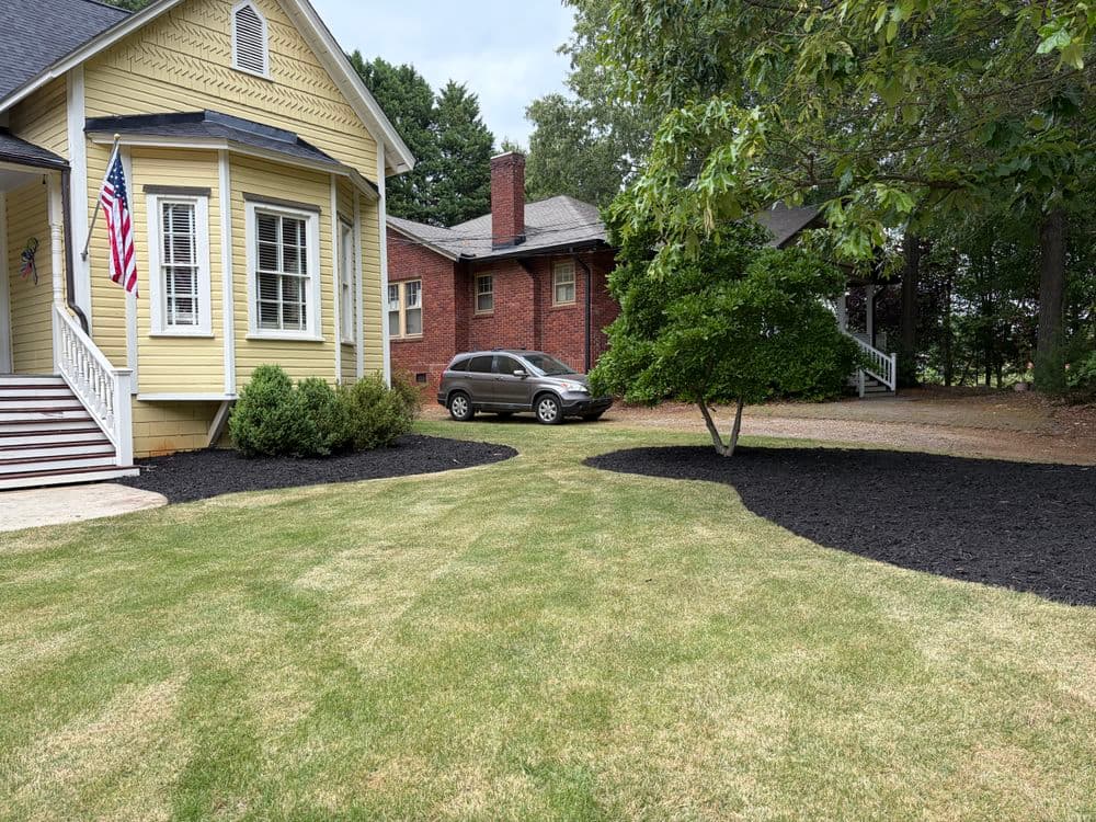 Yellow house with American flag, manicured lawn, and parked car on gravel driveway.